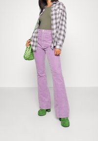 Pantalons évasés en velours lilas, chemise à carreaux gris avec boutons, débardeur vert olive, et sac en maille verte. Chaussures compensées vertes épaisses.