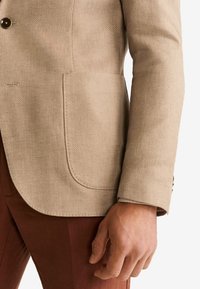 Béžový strukturovaný blazer s náprsní kapsou, nošený s hnědými kalhotami, ukazující dolní část trupu a pravou ruku osoby.