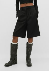 Pantalones cortos negros de corte entallado con cintura alta, pliegues y bolsillos laterales, combinados con botas de cuero negras hasta la rodilla con un detalle de hebilla.