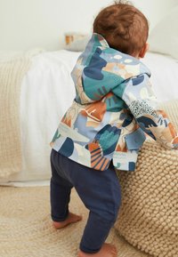 Giacca impermeabile colorata e a fantasia in toni di blu con forme di animali; abbinata a pantaloni blu scuro; superficie texturizzata. Bambino che allunga la mano verso un oggetto.