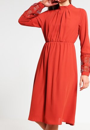 Femme portant une robe orange brûlé longueur genou avec taille froncée, col haut et détail en dentelle sur les manches longues.