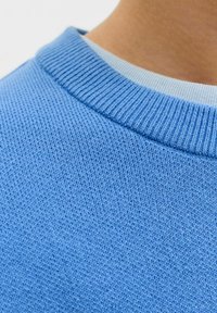 Gros plan sur un pull bleu clair à col rond porté par-dessus une chemise de couleur claire, montrant la texture tricotée et la zone du col au niveau du cou d'une personne.