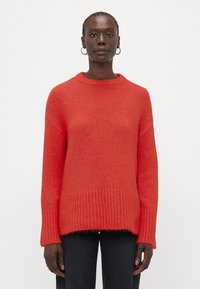 Maglione rosso lavorato a maglia con una finitura testurizzata, caratterizzato da un orlo a coste e maniche larghe, abbinato a jeans scuri.