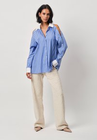 Blue striped shirt with cut-out shoulders, white collar, paired with cream textured wide-leg pants and pointed beige shoes.