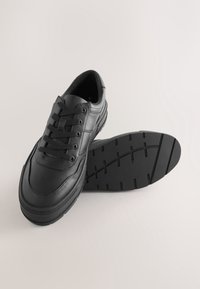 Paire de baskets en cuir noir avec lacets à l'avant et semelles épaisses en caoutchouc, une chaussure droite et l'autre montrant le motif de la semelle.