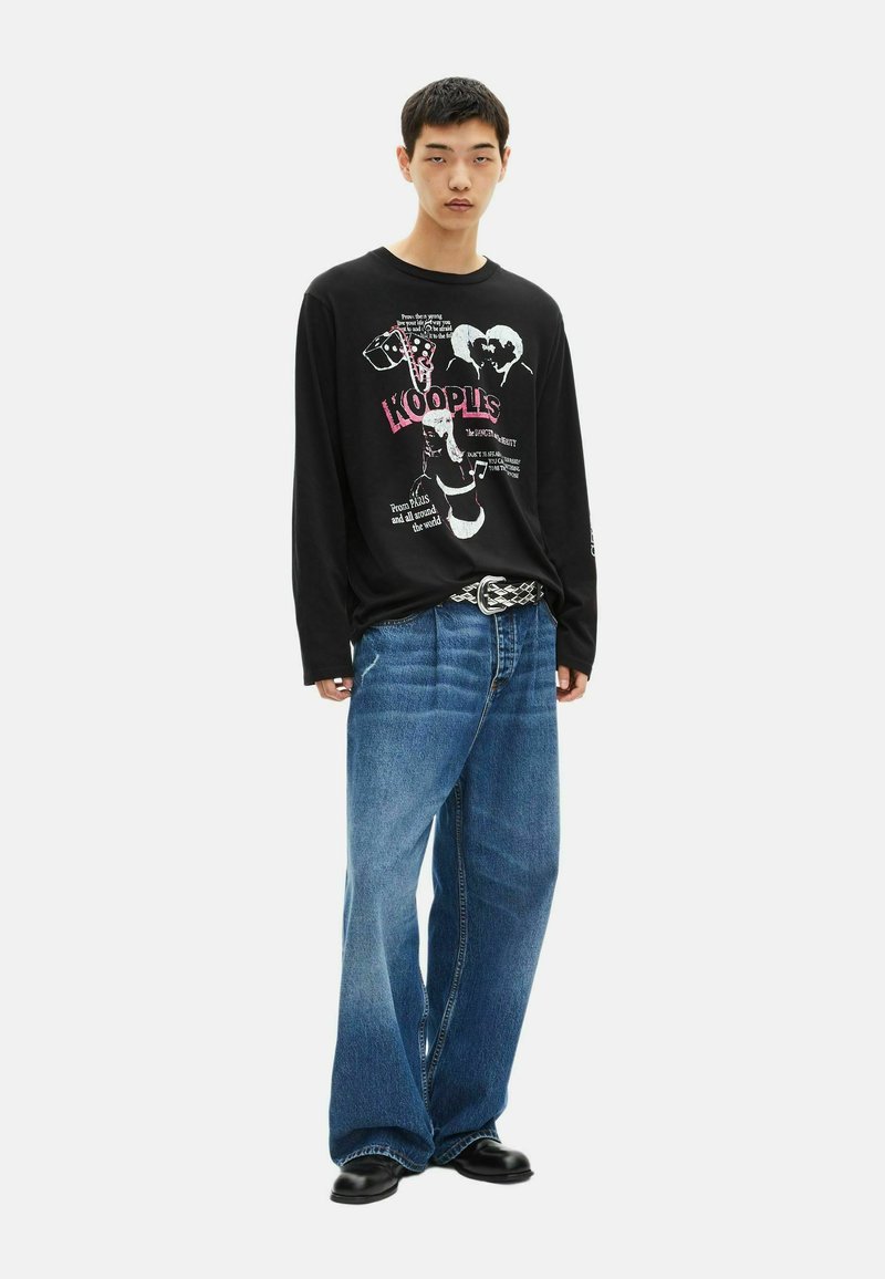 Giovane in camicia a maniche lunghe nera con grafica, jeans larghi blu, cintura a fantasia e scarpe nere, in piedi contro uno sfondo bianco.