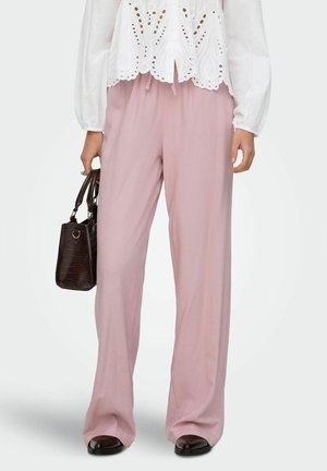 Femme portant un pantalon ample rose clair, un chemisier blanc à manches longues ajouré, tenant un sac à main sombre texturé, et des chaussures noires.