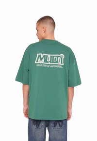 T-shirt verde oversize con maniche corte. Presenta un logo bianco e la scritta “MULTIPLY APPAREL” stampata sul retro. Texture liscia.