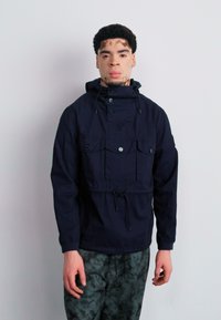 Navy pullover jacket with a high collar, front pockets, and drawstring waist. Material appears smooth, with a relaxed fit and functional design.