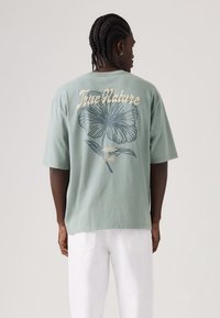 Πανίνο μπλουζάκι μέντας πράσινο με μεγάλο, περιγράμμινο ανθικό σχέδιο σε μπλε και το κείμενο «True Nature» σε κρεμ στο πίσω μέρος.