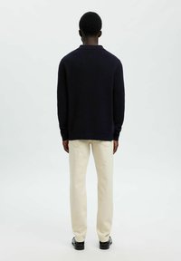 Dunkelblauer Strickpullover mit Kragen und langen Ärmeln, kombiniert mit hellbeigen, gerade geschnittenen Hosen und schwarzen Schuhen. Schlichter Hintergrund.