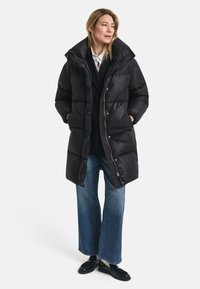 Zwarte pufferjas met een hoge kraag, voorzien van een ritsluiting en knoopaccenten. Draag deze over een zwarte trui en blauwe wijde jeans.