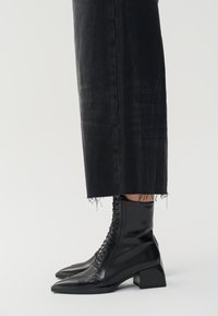 Bottines noires en cuir avec un bout pointu, un laçage à l'avant et un talon empilé, associées à un pantalon en denim foncé avec des ourlets effilochés.
