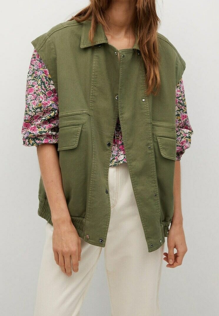 Femme portant un gilet sans manches vert olive boutonné par-dessus un chemisier rose à fleurs avec un pantalon crème, debout devant un fond clair uni.
