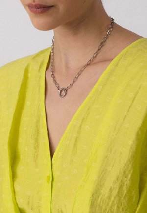 Frau trägt eine silberne Kettenhalskette mit einem runden Anhänger, kombiniert mit einem leuchtend gelben, strukturierten V-Ausschnitt-Top.