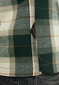 Gros plan sur l'ourlet d'une chemise à carreaux verts, beige et blancs avec des coutures visibles et une petite étiquette noire avec texte orange, portée sur un pantalon foncé.