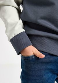 Ciemnoszara i jasnoszara bluza z prążkowanymi mankietami, noszona z niebieskimi dżinsami. Dłoń w kieszeni podkreśla swobodny krój i miękką fakturę tkaniny.