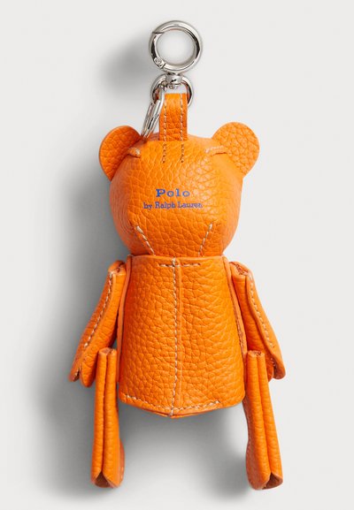 Kožený prívesok na kľúče v oranžovej farbe, tvarovaný ako medvedík s textúrovaným povrchom, s kovovým strieborným krúžkom a modrým logom „Polo by Ralph Lauren“.