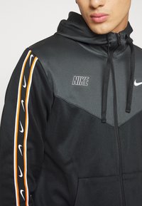 Svart zip-up hoodie i slätt tyg med huva, med ett chevrondesign och orangea och vita Nike-logotyper längs ärmarna.