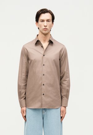 Άνδρας στέκεται φορώντας πουκάμισο σε σκούρο μπεζ χρώμα με κουμπιά και μακρύ μανίκι και ανοιχτό μπλε τζιν, μπροστά σε απλό λευκό φόντο.