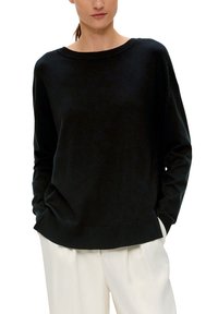 Schwarzer gestrickter Pullover mit weiter Halslinie, fallenden Schultern und gerippten Bündchen. Kombiniert mit weißen, locker sitzenden Hosen.