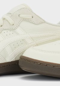 Gros plan d'une chaussure de sport blanche avec un panneau à œillets en daim beige, des côtés en cuir perforé et une semelle en caoutchouc marron.