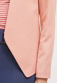 Broskvový blejzr s hladkou texturou, s ostrým límcem a zkoseným předním lemem, zkombinovaný s pruhovaným topem uvnitř a námořnickými kalhotami.