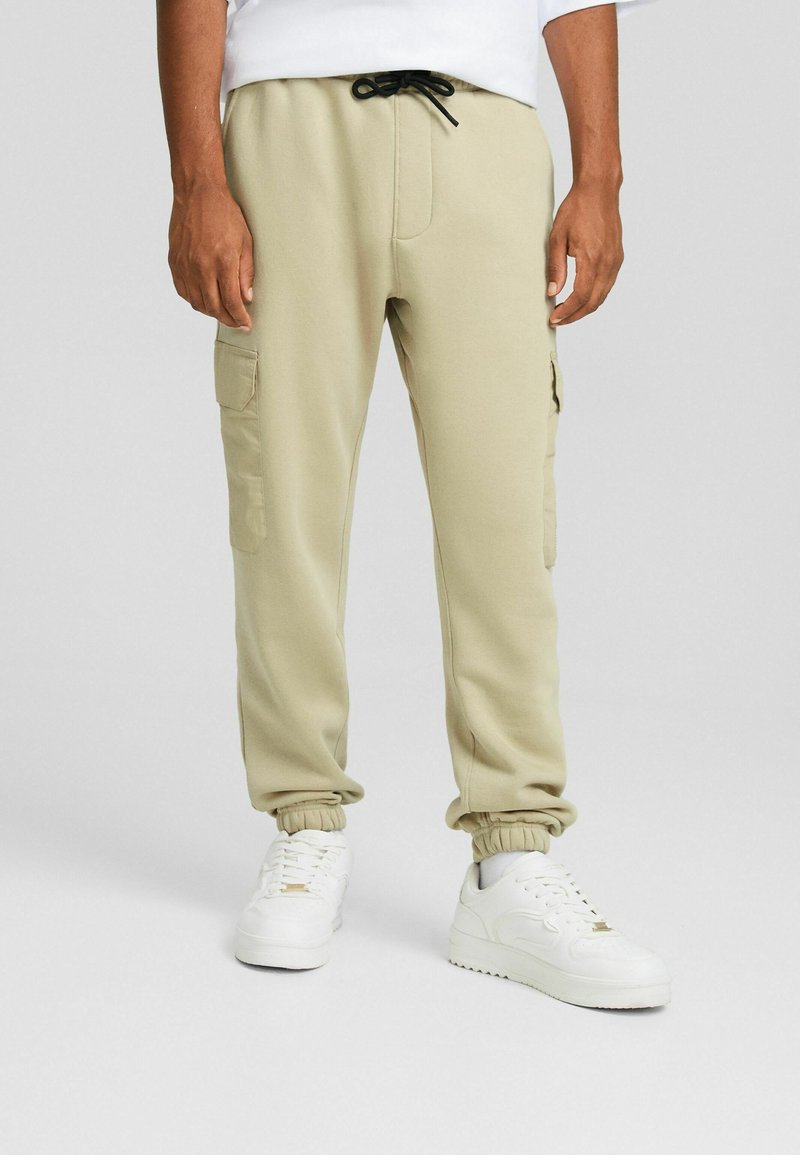 Beige cargobyxor med elastisk midja och fållar. Har sidofickor och dragsko. Bärs med vita sneakers.