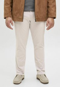 Pantalons beige clair en tissu doux, design fuselé, associés à des chaussures slip-on beiges. Veste marron avec une texture lisse.