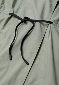 Cordon noir noué en nœud sur un tissu vert olive clair avec des coutures visibles et une texture légèrement froissée.