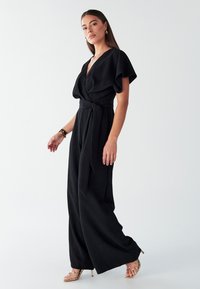 Črni kombinezon z globokim V-izrezom, učinkovito padajočimi rokavi in pasom, zavezanimi v pentljo. Ponaša se s širokimi hlačami iz gladkega blaga. Obut z zlatimi sandali na tanko peto.