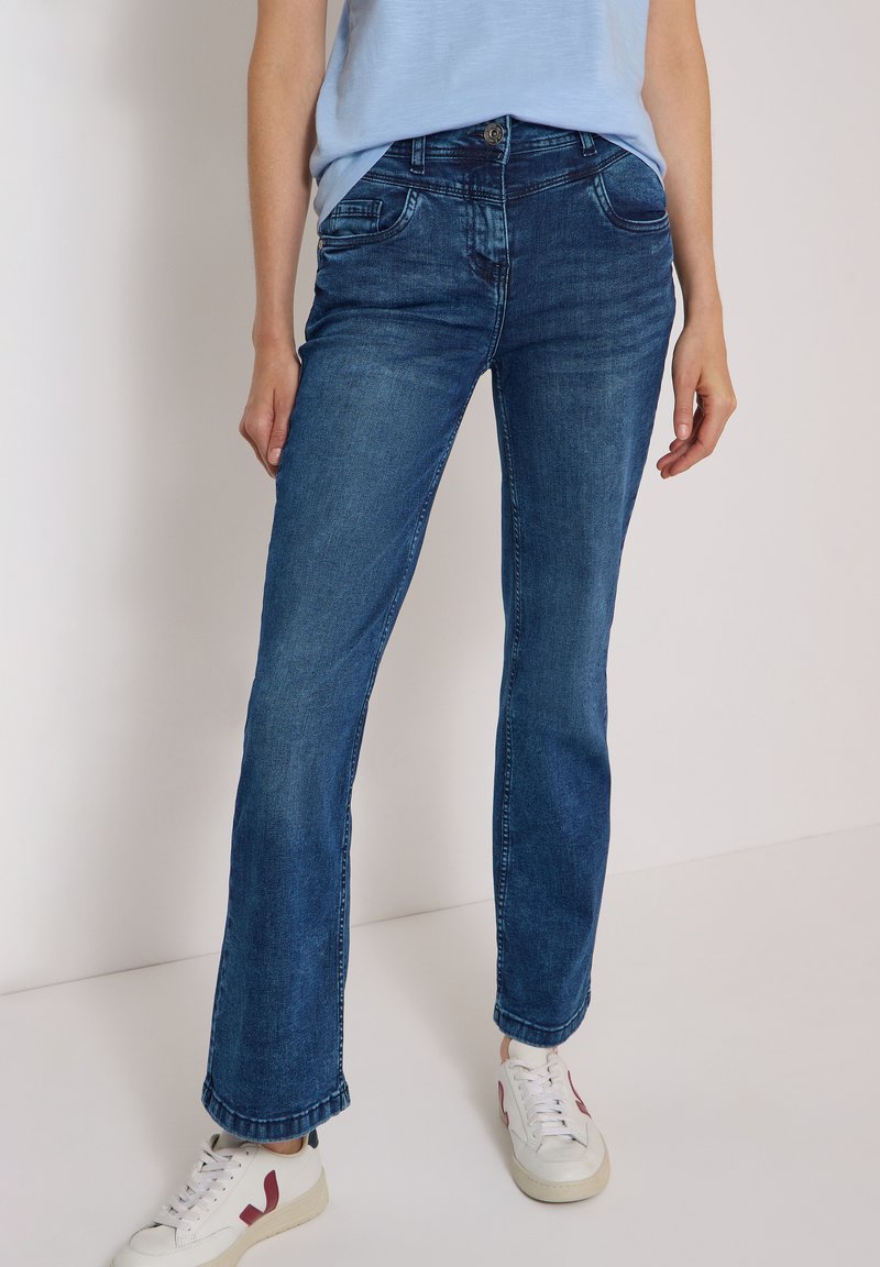 Personne portant un jean bleu foncé droit taille mi-haute, un haut bleu clair et des baskets blanches avec des touches rouges, debout devant un fond uni.
