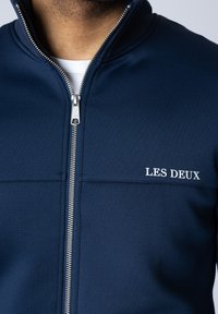 Jachetă navy cu fermoar, dintr-un material neted, având un guler înalt, fermoar argintiu și logo-ul "LES DEUX" în alb pe pieptul stâng.