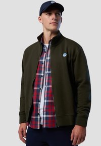 Giacca verde scuro con zip e colletto alto, indossata sopra una camicia a scacchi rossa, bianca e blu. Presenta una toppa con logo sul lato sinistro del petto.
