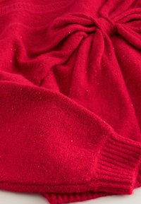 Red knitted sweater featuring a textured finish, with a bow detail at the front and ribbed cuffs. Soft fabric with subtle flecks.