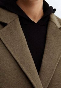 Manteau en laine vert olive avec un col cranté, porté par-dessus un sweat à capuche noir texturé. Vue en gros plan mettant en valeur les détails du tissu et de la conception.