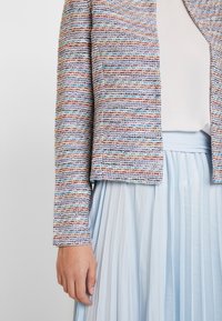 Blazer court texturé et multicolore avec un motif tissé, assorti d'une jupe plissée bleu clair. Manches longues et design ouvert à l'avant.