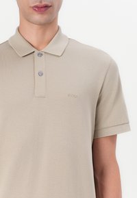 Bežica polo majica s teksturiranim materijalom, kratkim rukavima i ovratnikom. Ima dugmasti izrez i diskretno izvezenu oznaku na prsima.