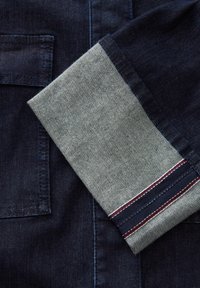 Tessuto di denim scuro con un risvolto grigio chiaro a contrasto, caratterizzato da un bordo a strisce in rosso, bianco e blu, che mostra un design pulito e sartoriale.