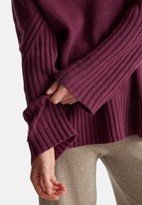 Strickpullover in tiefem Lila mit Rippstruktur an den Ärmeln. Die Hand des Models hält den Ärmel und zeigt den Stoffmix sowie das lässige Design.