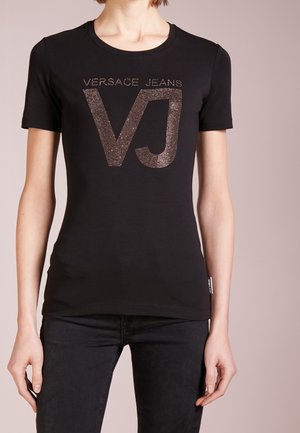 Czarna dopasowana koszulka z krótkimi rękawami i złotym, przytwierdzonym logo "VERSACE JEANS VJ" z przodu, noszona z czarnymi spodniami.