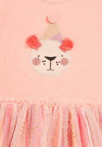 Vaaleanpunainen t-paita, jossa on kirjailtu karhun kasvot juhlapäähineellä. Rypistetty nylonhame vaaleanpunaisena, kullankirjavilla täplillä lisää tekstuuria ja yksityiskohtia.