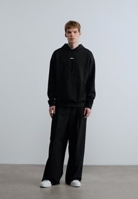 Μαύρο φούτερ με κουκούλα και κορδόνια, με λογότυπο, συνδυασμένο με μαύρο παντελόνι φαρδιάς γραμμής. Το σύνολο ολοκληρώνεται με λευκά αθλητικά παπούτσια, με φόντο ουδέτερο χρώμα.