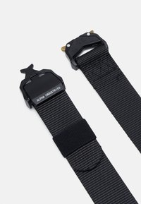 Ceinture noire en nylon avec tissage texturé, dotée de deux boucles en métal noir ; l'une possède des accents dorés et un logo en relief. Fermeture à crochet et boucle incluse.