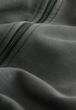 Szürke anyag sima textúrával, amelyen zöld cipzár található. Az anyag finom kötésmintát mutat, kiemelve a textília tartósságát és rugalmasságát.