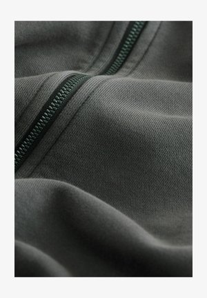 Szürke anyag sima textúrával, amelyen zöld cipzár található. Az anyag finom kötésmintát mutat, kiemelve a textília tartósságát és rugalmasságát.