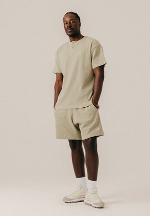 Mann mit geflochtenem Haar steht mit den Händen in den Taschen, trägt ein beige Kurzarmhemd, passende Shorts, weiße Socken und Sneakers vor hellem Hintergrund.