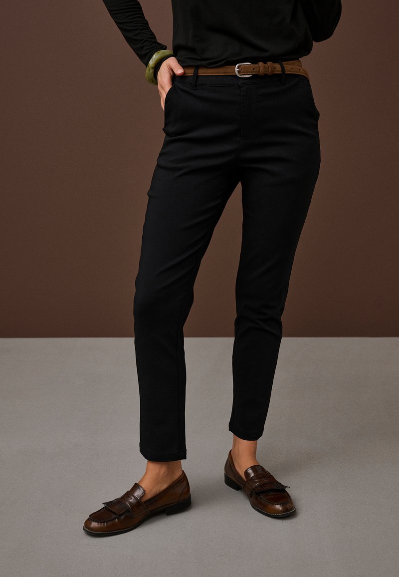 Osoba nosící černé úzké kalhoty, hnědý kožený pásek, hnědé mokasíny a černý dlouhý rukáv, s jednou rukou v kapse na jednoduchém pozadí.