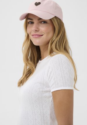 Tânără cu păr lung și blond, purtând o șapcă roz cu o inimă și un top alb cu mâneci scurte și model, zâmbind ușor, orientată spre stânga.