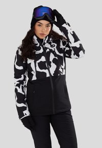 Frau, die eine schwarz-weiß gemusterte Skijacke, schwarze Hose, Handschuhe, Mütze und eine Skibrille mit blauer Tönung trägt und mit einer Hand die Skibrille justiert.